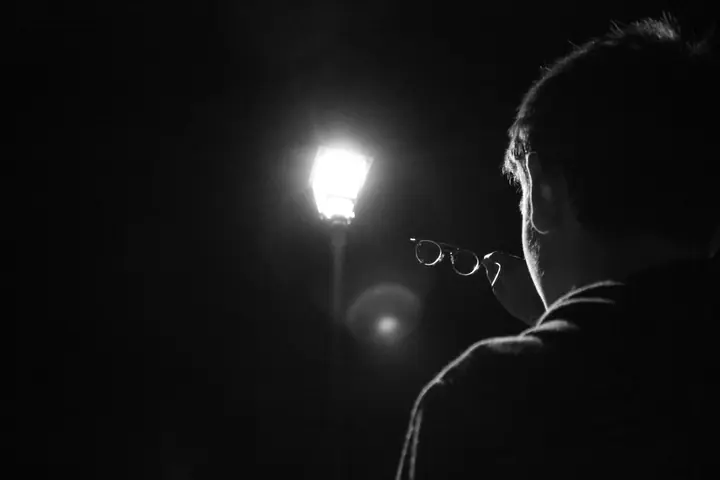 Un homme de dos, ses lunettes à la main, face à un réverbère allumé dans la nuit