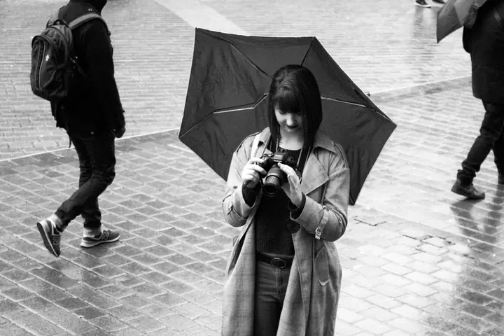 Une jeune femme sous un parapluie règle son appareil photo, sur une place pavée sous la pluie