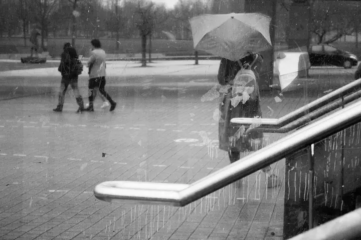 Une place sous la pluie vue à travers une vitre, avec des passants et un parapluie