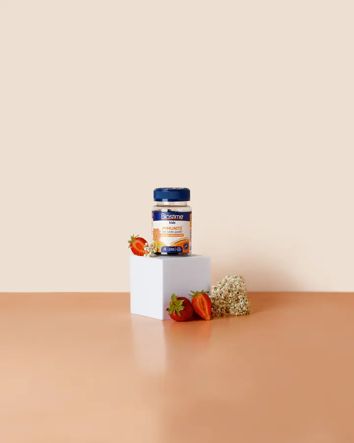 Un pilulier de gommes Biostime sur un cube blanc, entouré de fruits rouges et de fleurs, sur fond crème