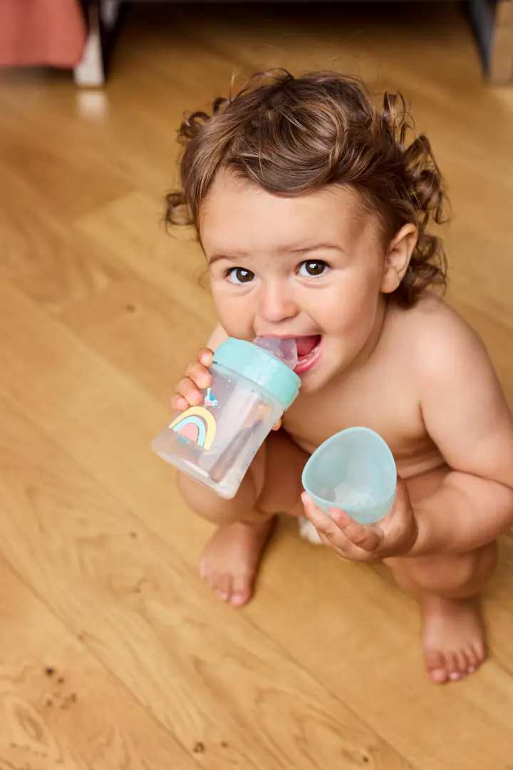 Un bébé boit dans un gobelet d’apprentissage, assis sur un parquet