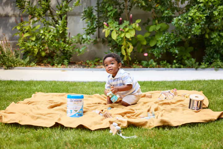 Un jeune enfant joue sur un plaid jaune dans l’herbe, près d’une boîte de lait Biostime