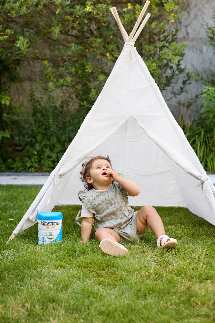Un bébé assis devant un tipi blanc dans un jardin, à côté d’une boîte de lait Biostime