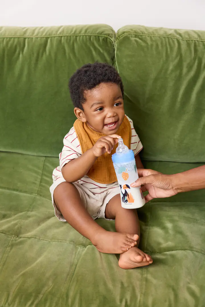 Un jeune enfant souriant sur un canapé vert, un biberon à la main