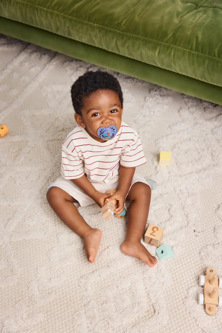 Un jeune enfant à la tétine, assis sur un tapis avec des jouets en bois
