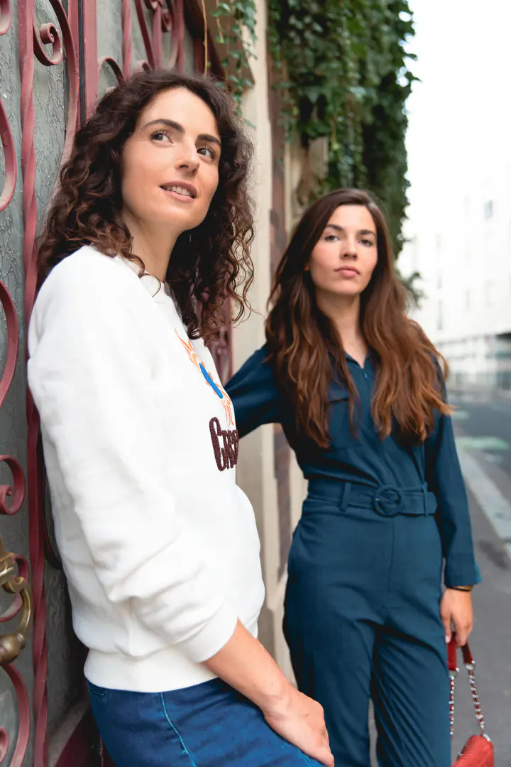 Deux femmes adossées à une porte en fer forgé, l’une en sweat blanc, l’autre en combinaison en jean